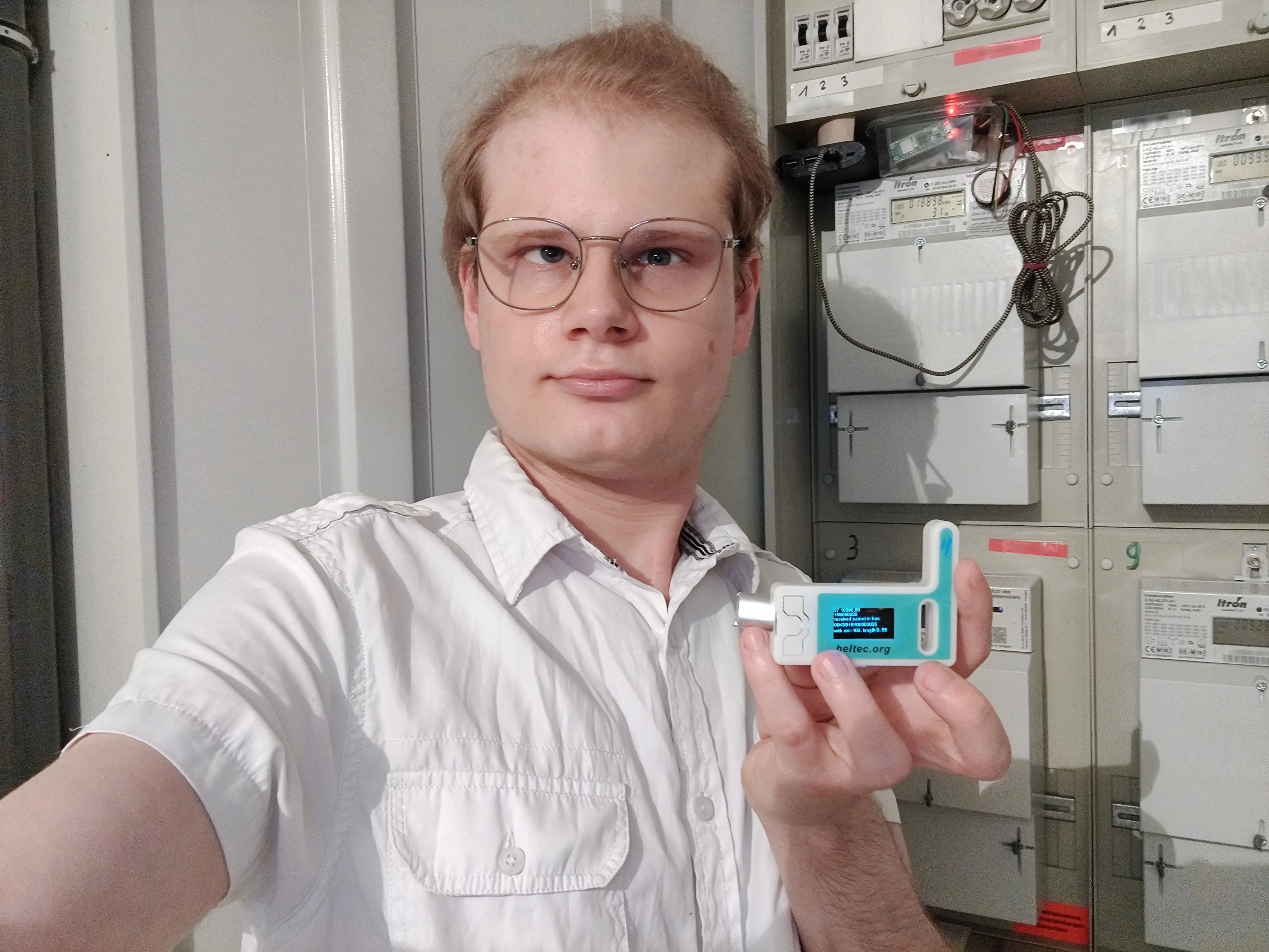 Vorschaubild Stromzähler HQ, Janis hält einen ESP32 in der Hand von dem Display den Stromverbrauch anzeigt. Im Hintergrund sind Stromzähler, an einem hängt ein Pico W mit Lesekopf